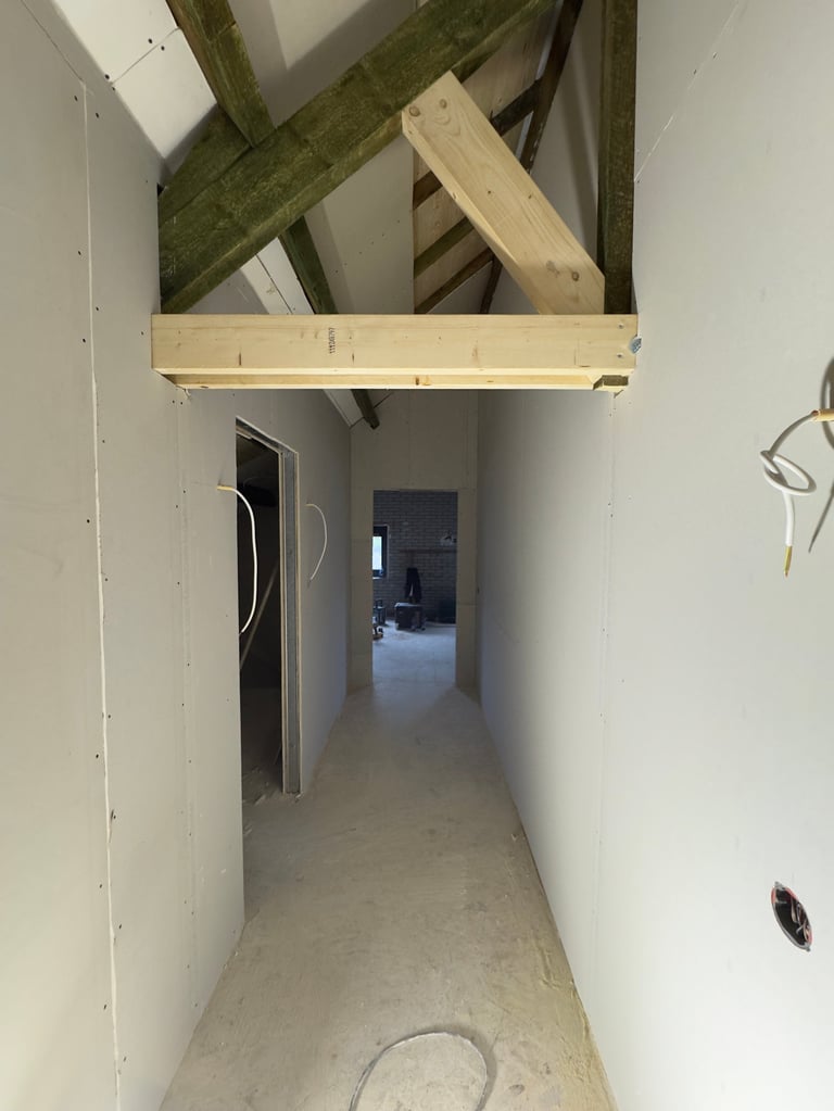 Narrow hallway under construction with exposed wooden beams, drywall on walls, and concrete floor leading to an open doorway