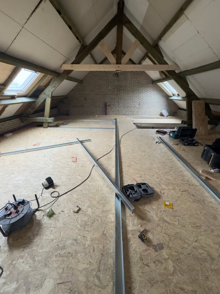 Unfinished attic space under construction with exposed wooden beams, skylights, brick wall, and construction materials scattered on plywood flooring