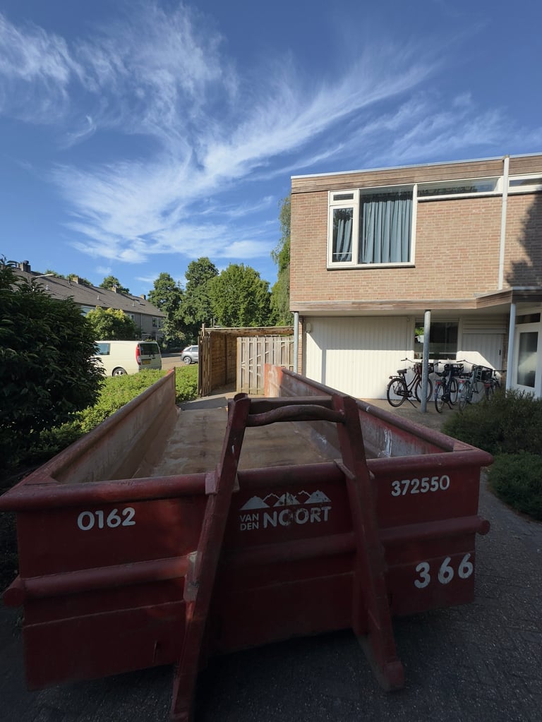 Red metal dumpster or waste container in driveway with brick house, garage, and blue sky in background