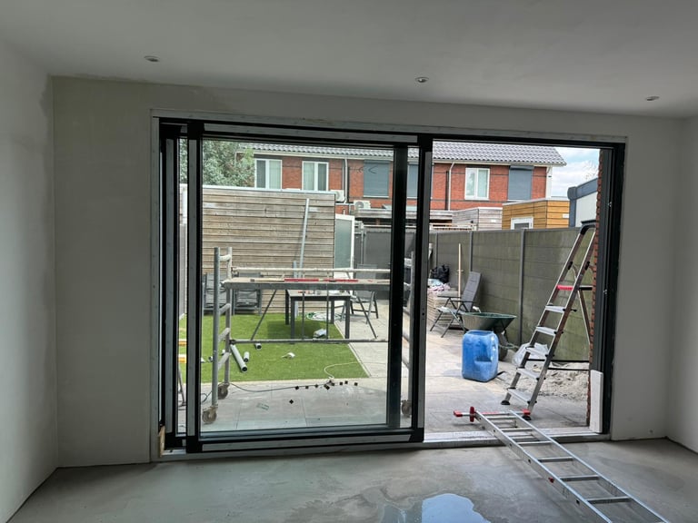 View through open black bi-fold doors from indoors to a construction site with green lawn, brick buildings, and tools visible outside