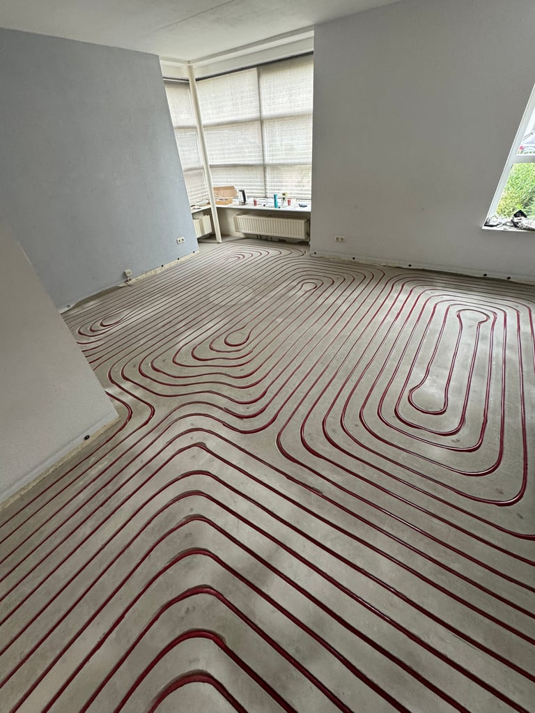 Empty room with radiant floor heating system installed, showing red tubing in serpentine pattern on concrete floor with white walls and windows