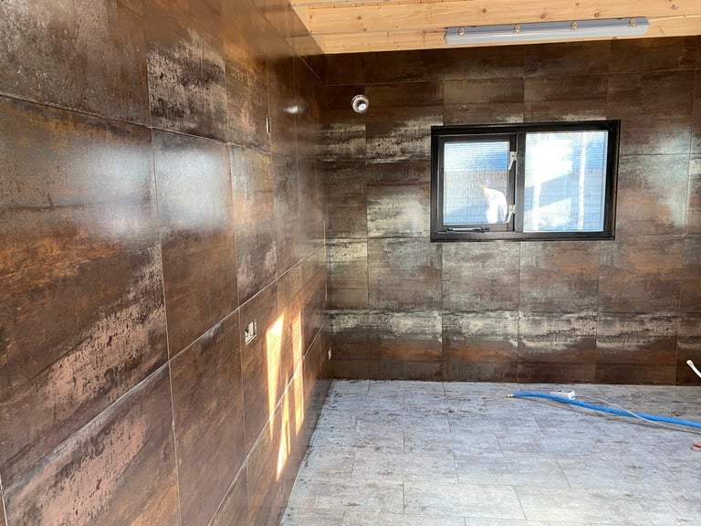 Empty room under construction with brown tile walls, wooden ceiling, two windows, and blue hose on concrete floor