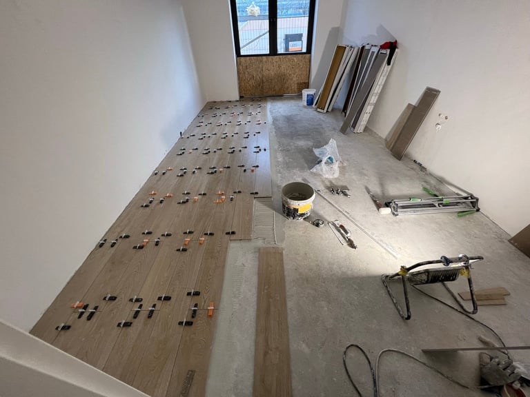 Interior construction site with wood flooring installation in progress, tools scattered on floor, and building materials stacked against walls