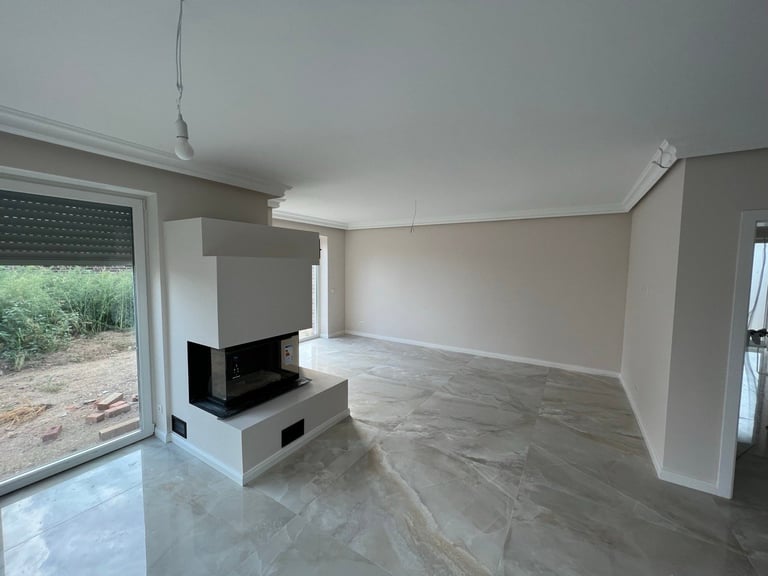 Modern empty living room with polished concrete floors, white walls, central fireplace, and large glass doors leading to outdoor garden area